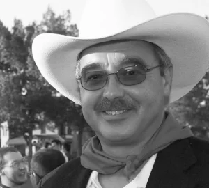 A man with a mustache and glasses wears a cowboy hat and a neckerchief.