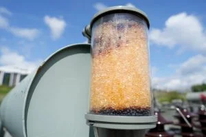 A clear cylindrical container is filled with small orange and dark beads.