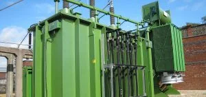 A large green electrical transformer stands outdoors next to a brick wall.