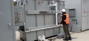 A person in a white hard hat and orange safety vest stands next to a large piece of grey industrial machinery.