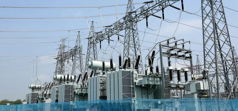 Electrical transformers and power lines stand within an electrical substation under a clear sky.