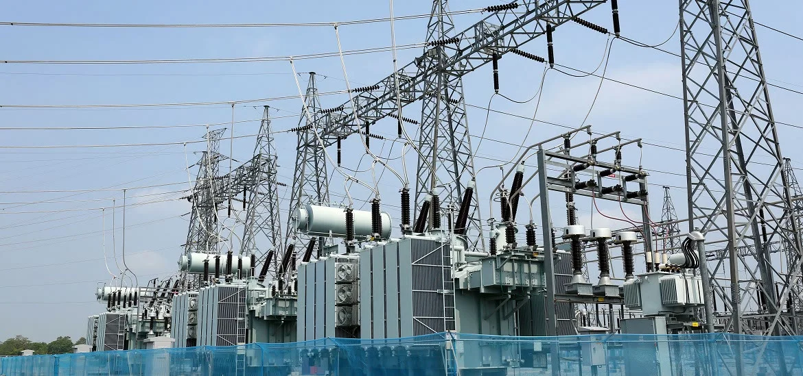 Electrical transformers and power lines stand within an electrical substation under a clear sky.