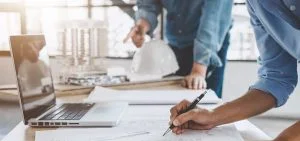 A person holds a pen over architectural blueprints on a desk next to a laptop and a model.