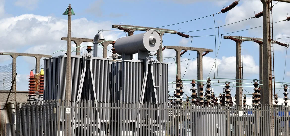 Large electrical transformers and power lines stand behind a metal fence under a cloudy sky.