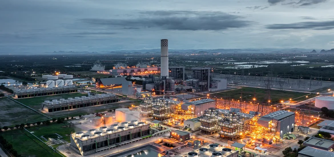 An industrial power plant is lit up at dusk under a cloudy sky.