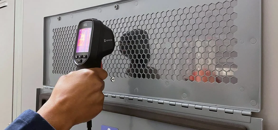 A hand holds a digital thermometer up to a perforated metal panel.