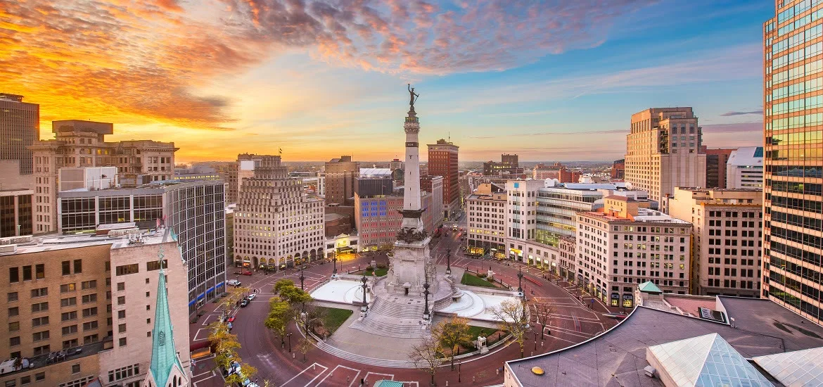 A tall monument stands in the center of a city square surrounded by buildings under a sunset sky.
