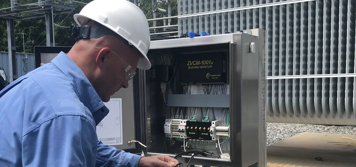 A person in a hard hat and blue shirt looks into an open electrical control box.