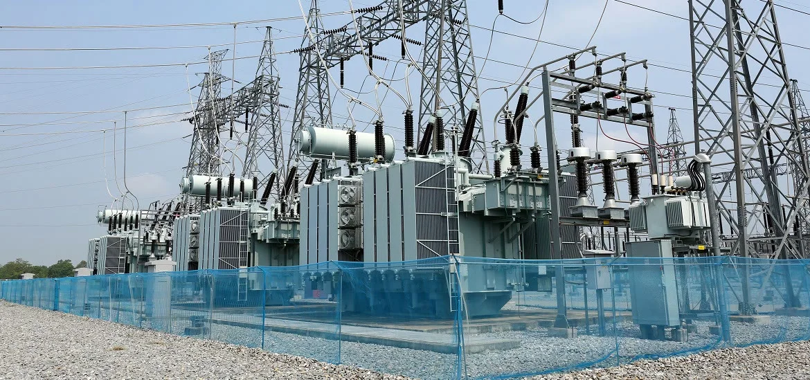 Electrical transformers and power lines stand behind a blue mesh fence.