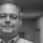 A man with glasses looks toward the camera in a black and white portrait.
