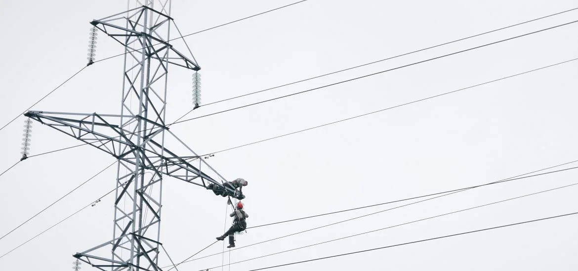 A worker in a red helmet hangs from a power line attached to a metal utility tower.