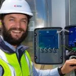A smiling man in a hard hat and safety vest holds an open electrical control box.