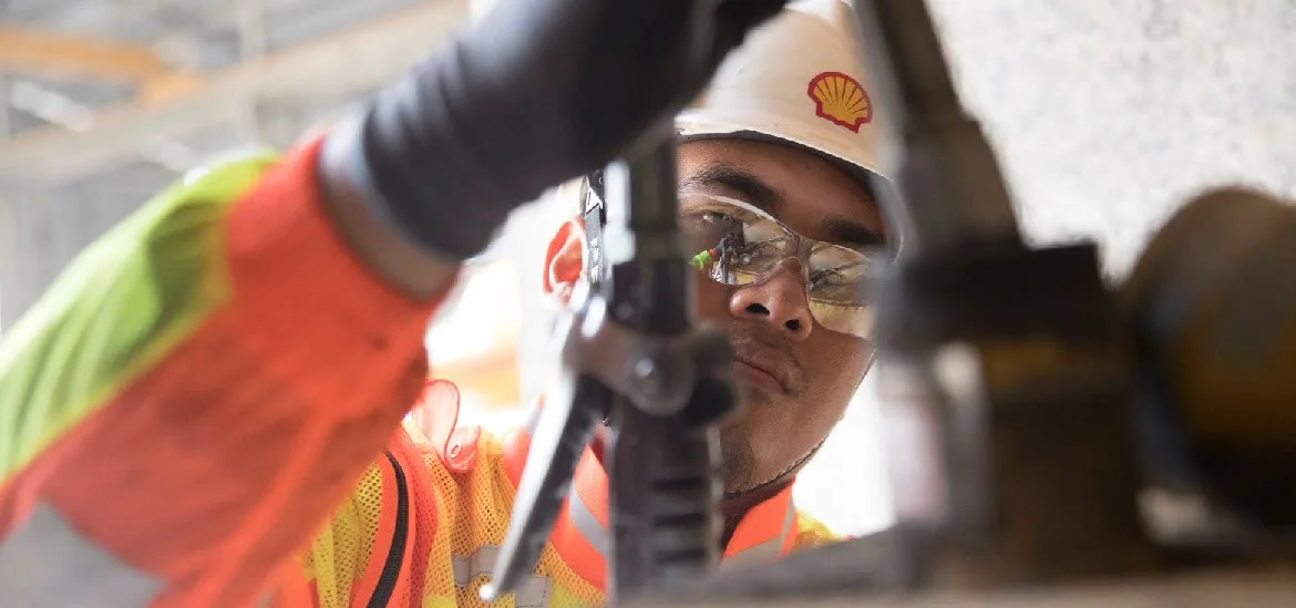 A person wearing a hard hat and safety glasses looks through a piece of machinery.