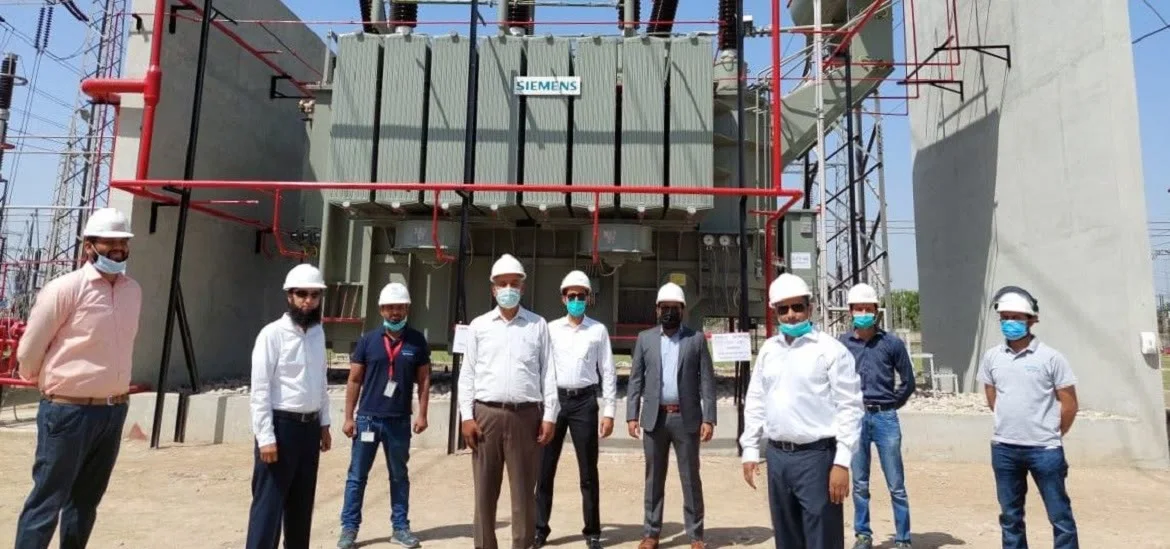 Seven men in hard hats and work clothes stand in a line in front of a large electrical transformer.