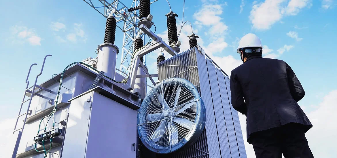 A person in a suit and hard hat stands next to a large electrical transformer.