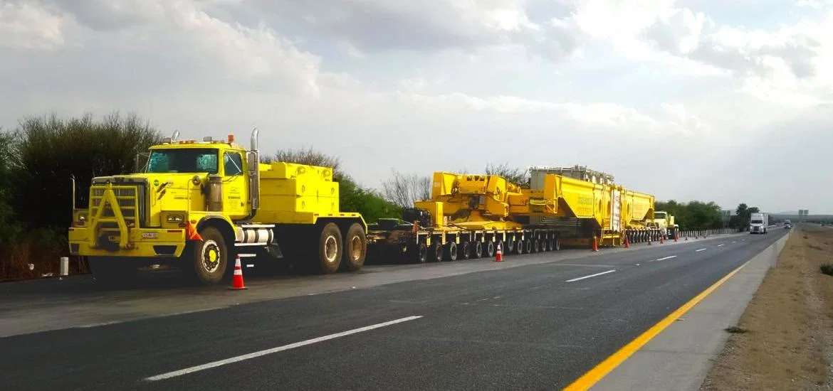 A yellow semi-truck pulls a long trailer carrying heavy construction equipment down a highway.
