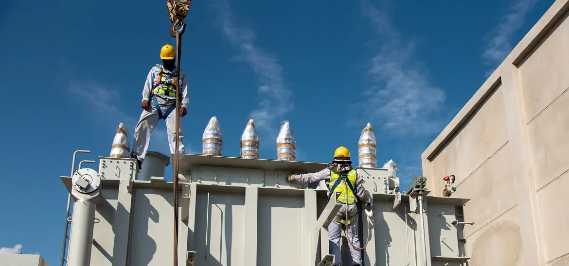 Two workers in hard hats and safety harnesses stand on top of a large industrial machine under a blue sky.