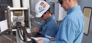 Two men in blue shirts work with a laptop and an open electrical control box.
