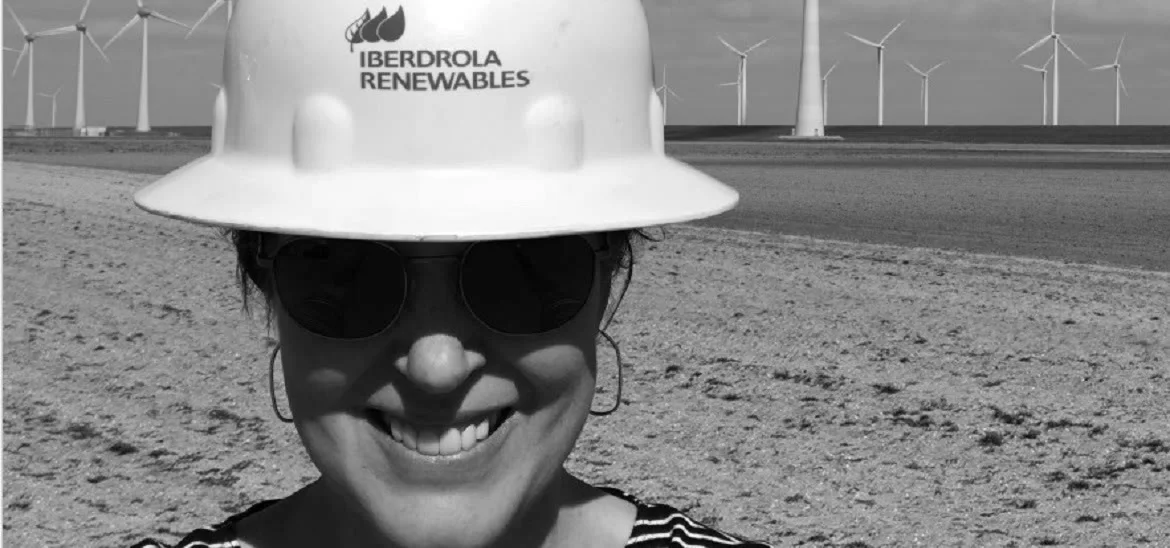 A person wearing a white hard hat and sunglasses smiles in front of wind turbines.