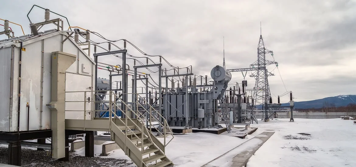 Electrical substation equipment sits in a snowy landscape under a cloudy sky.
