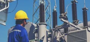 A person in a yellow hard hat and blue shirt looks toward electrical equipment.
