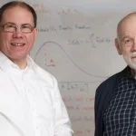 Two men stand in front of a whiteboard covered in mathematical equations.