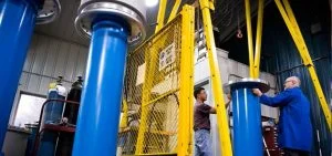 Two men stand near large blue cylinders and a yellow metal cage in a workshop.