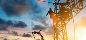 A silhouette of a person working on a power line tower against a sunset sky.