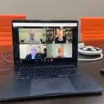 A laptop on a table displays a video call with four people.
