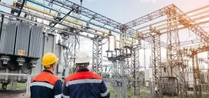 Two workers in hard hats and safety vests stand before an electrical substation.