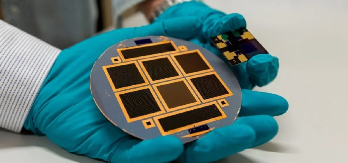 A person wearing blue gloves holds a circular silicon wafer with rectangular patterns.
