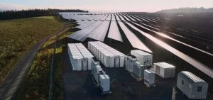 Rows of solar panels sit next to several white utility containers and a dirt path.