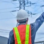 A person in a white hard hat and orange safety vest points toward power lines.