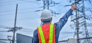 A person in a white hard hat and orange safety vest points toward power lines.