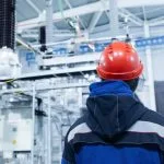 A person in a blue jacket and red hard hat looks toward industrial electrical equipment.