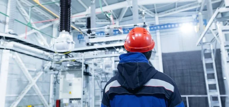 A person in a blue jacket and red hard hat looks toward industrial electrical equipment.