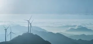 Wind turbines stand on misty mountains under a pale sky.