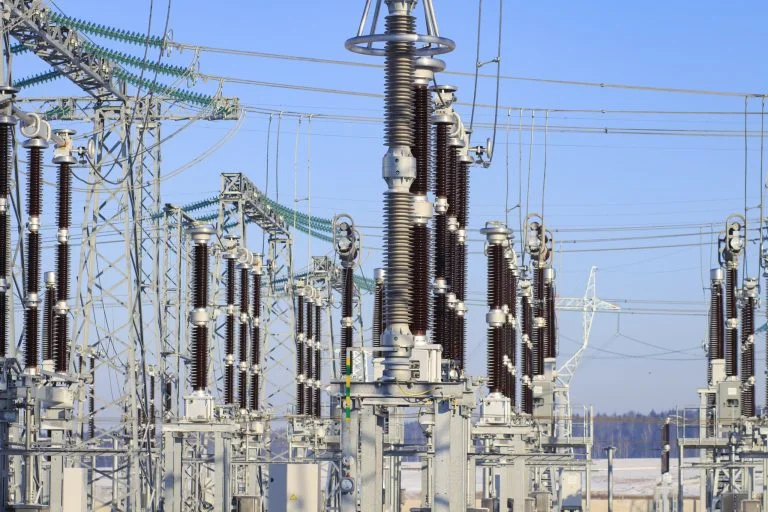 Electrical insulators and metal structures stand in an electrical substation under a clear blue sky.