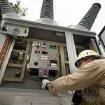 A person in a hard hat works on an open electrical control panel.