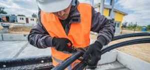 A person in a hard hat and orange safety vest uses a tool on black cables.