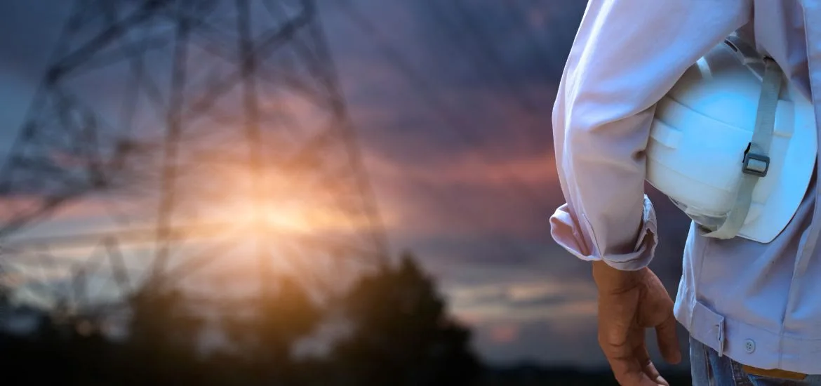 A person in a white shirt holds a white hard hat against a sunset background with a power line tower.
