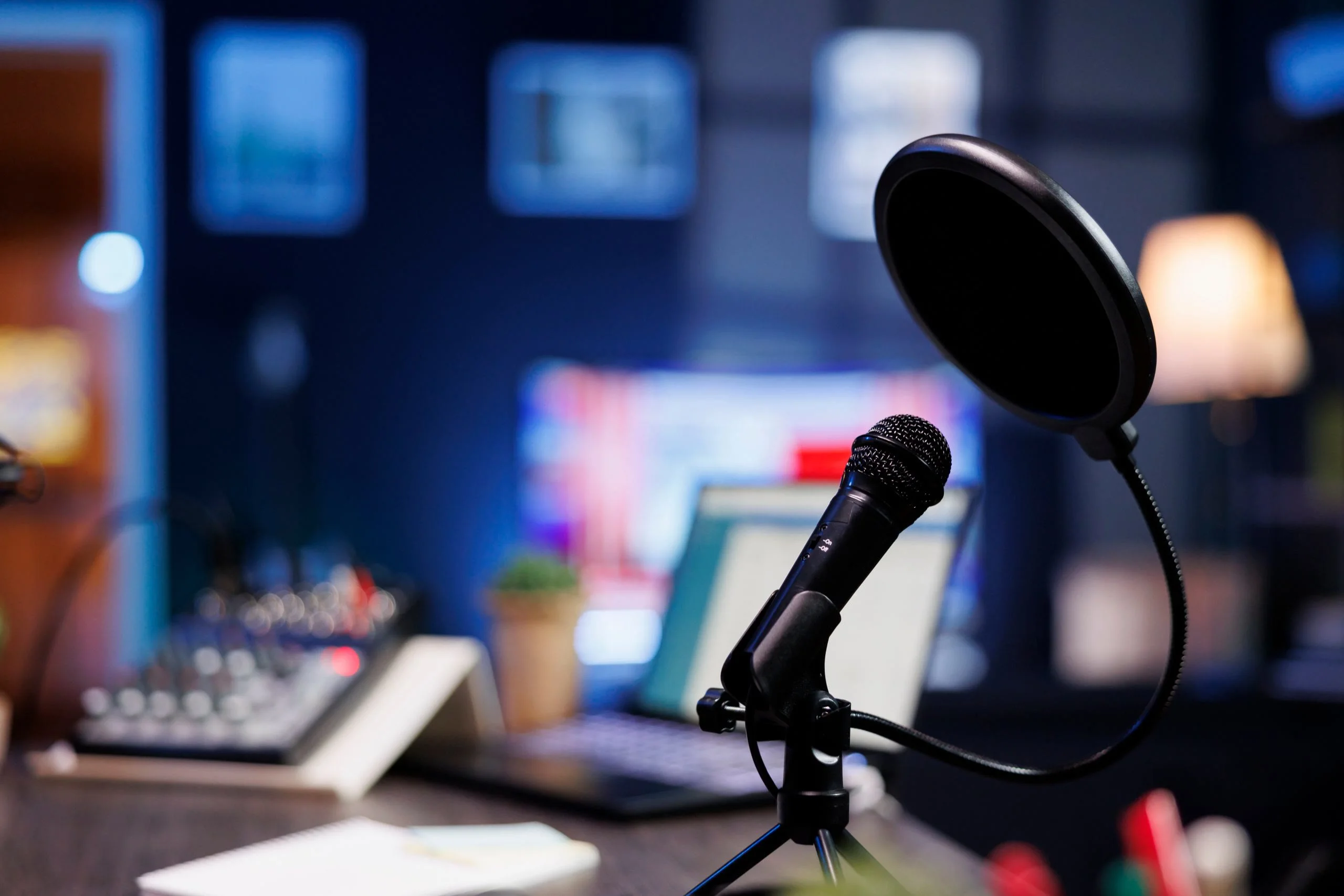 A microphone and pop filter sit on a desk in front of a blurred computer monitor.
